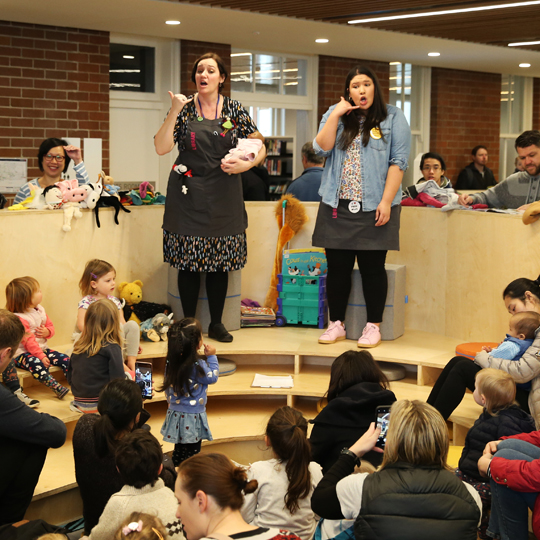 Two people stand above a crowd of families animatedly telling a children's story within a brightly lit library space